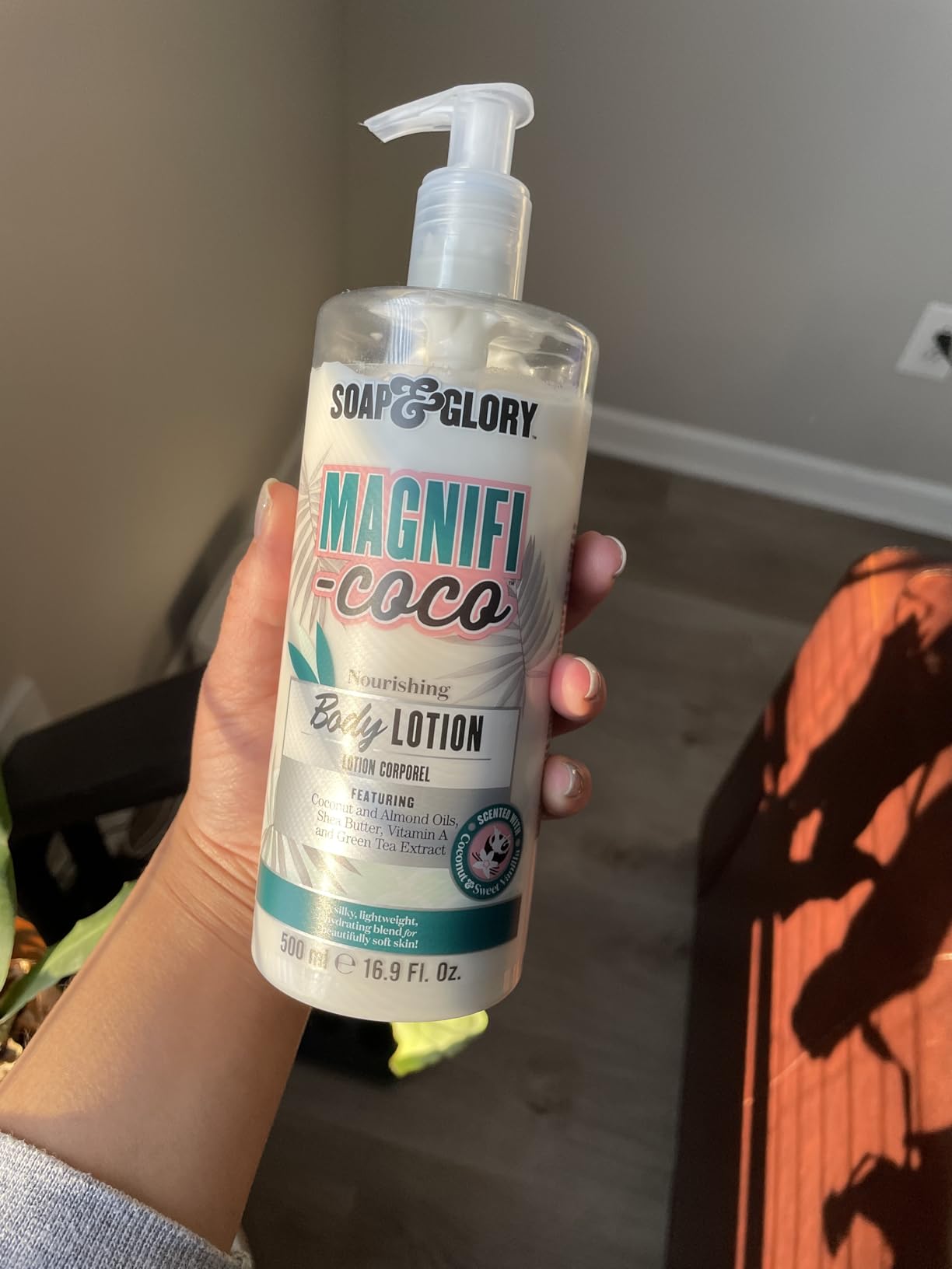 A hand holding a 500 ml Soap & Glory Magnifi-Coco Body Lotion pump bottle in front of a wood floor