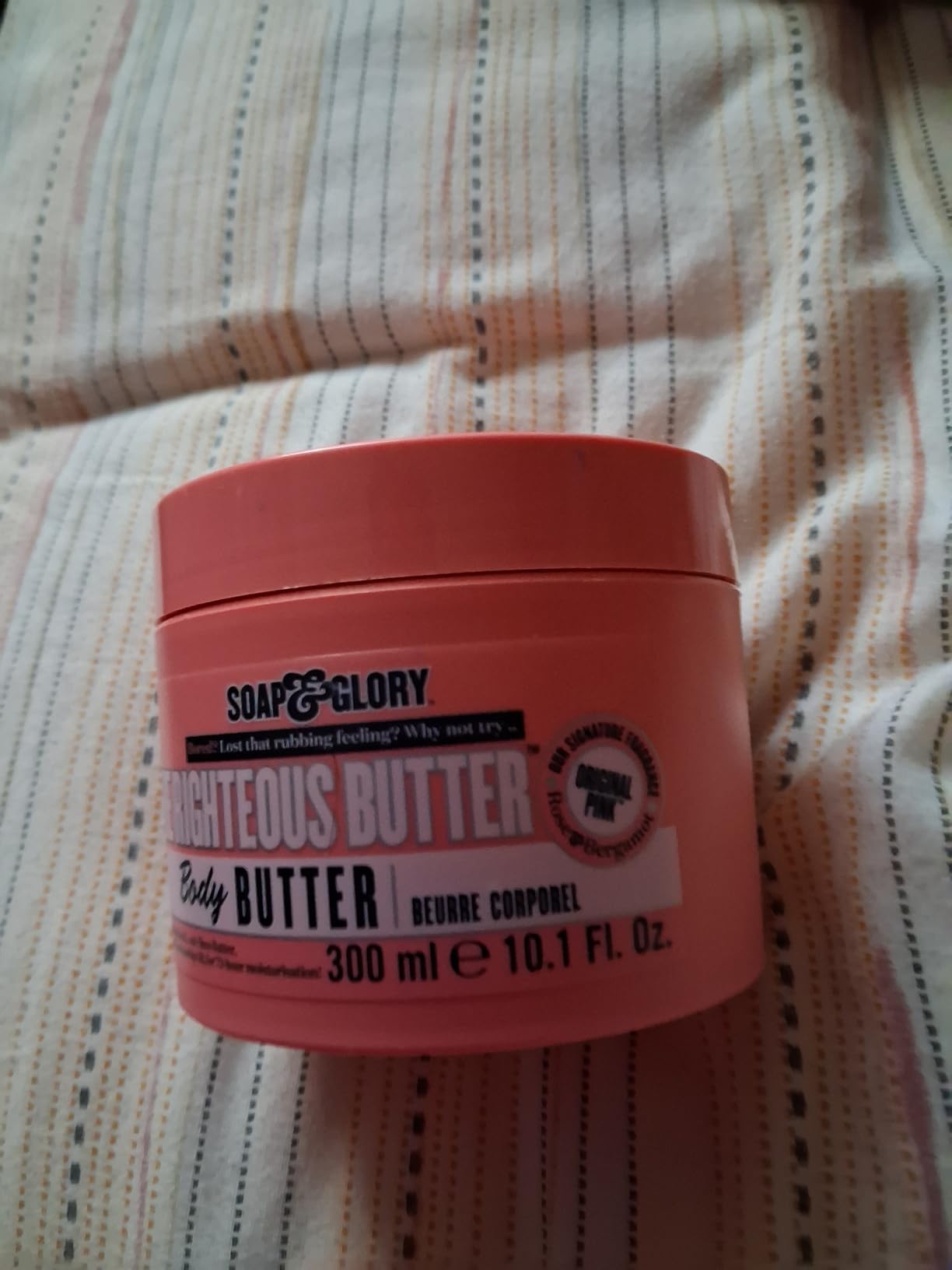 A 300 ml coral tub of Soap & Glory The Righteous Butter body butter resting on striped bedding