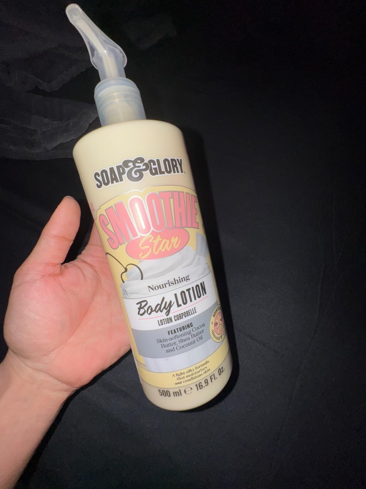 A hand holding a 500 ml Soap & Glory Smoothie Star Body Lotion pump bottle in low light